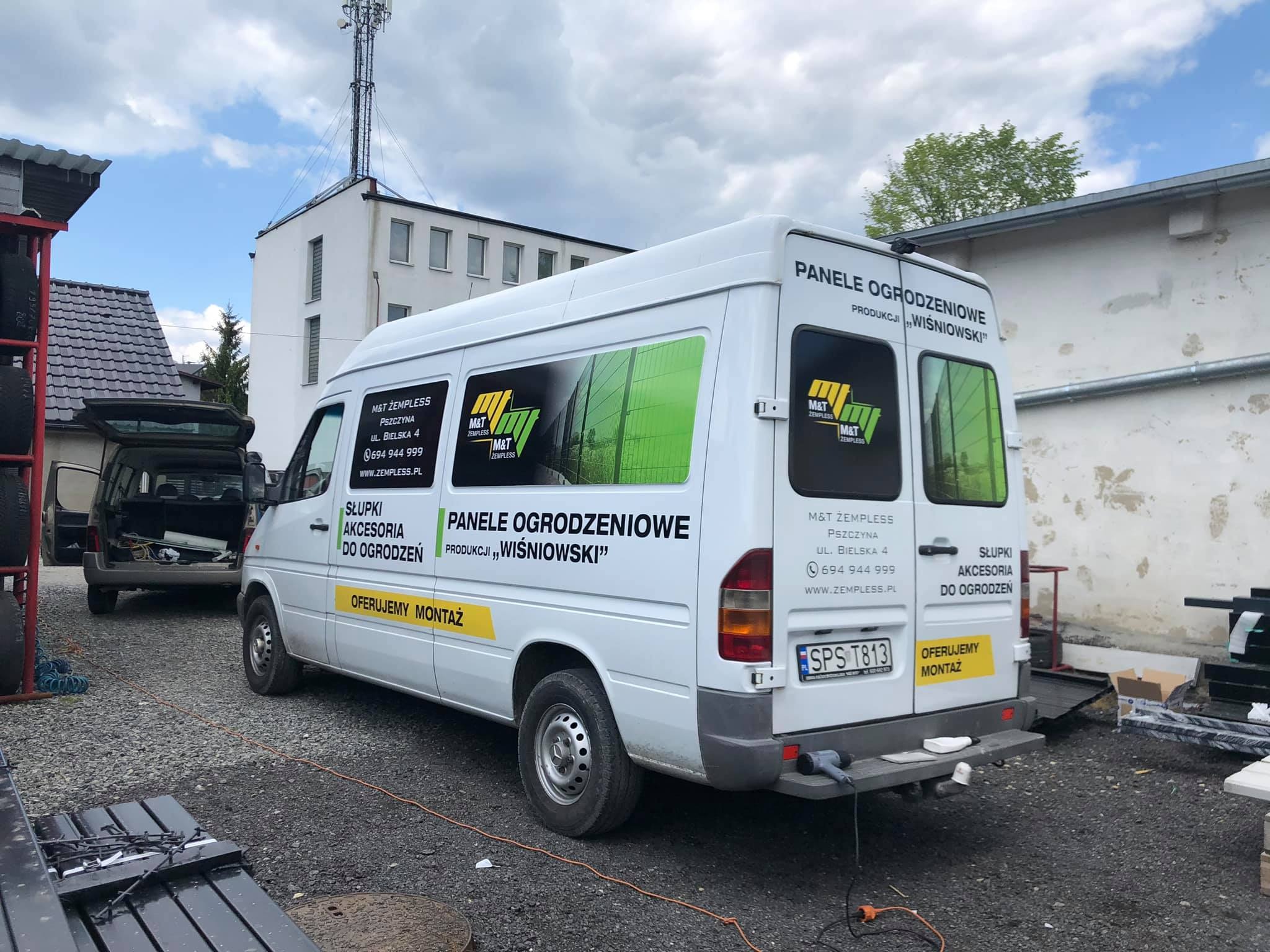 Biały samochód dostawczy z reklamą paneli ogrodzeniowych firmy 'Wiśniowski' na tle placu budowy z widocznymi elementami ogrodzeń.