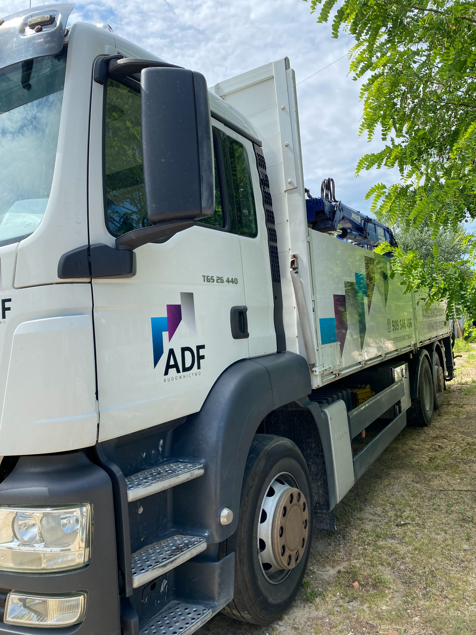 Biała ciężarówka MAN TGS 26.440 z logo ADF Budownictwo, widoczna z boku, z zamontowanym dźwigiem, zaparkowana na trawiastym terenie.