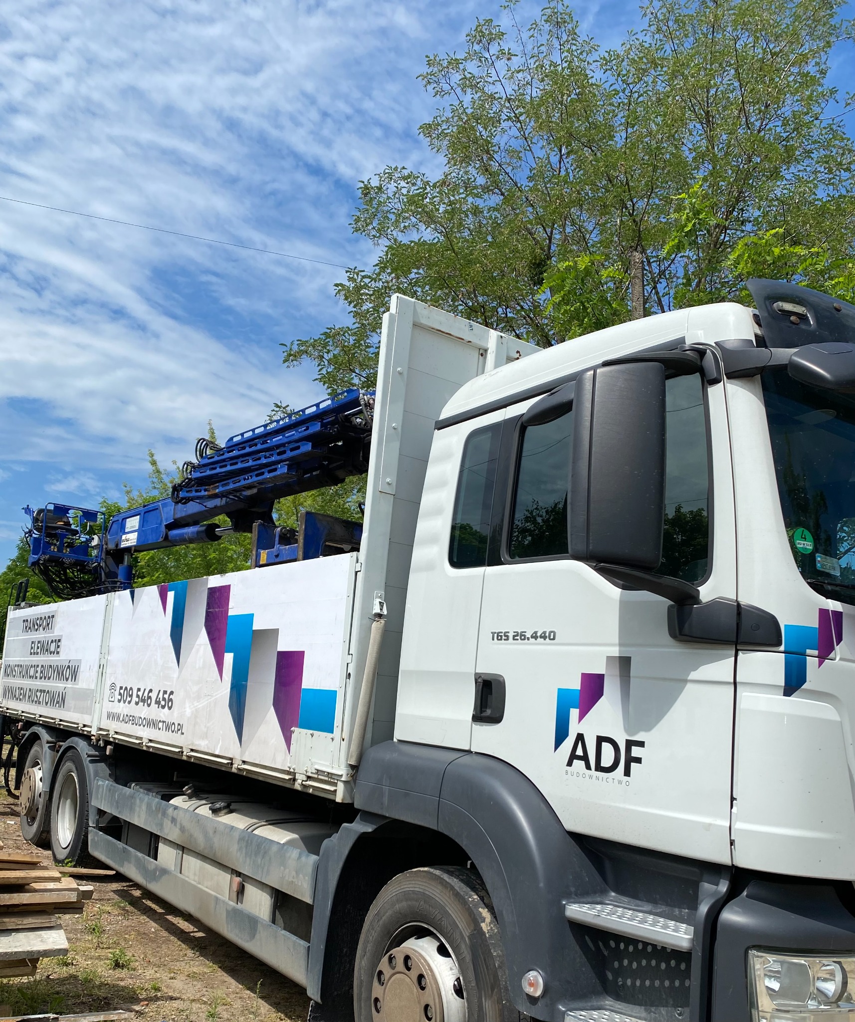 Ciężarówka MAN TGS 26.440 z niebieskim dźwigiem hydraulicznym na platformie, z reklamą firmy budowlanej ADF Budownictwo na boku, zaparkowana na placu budowy.