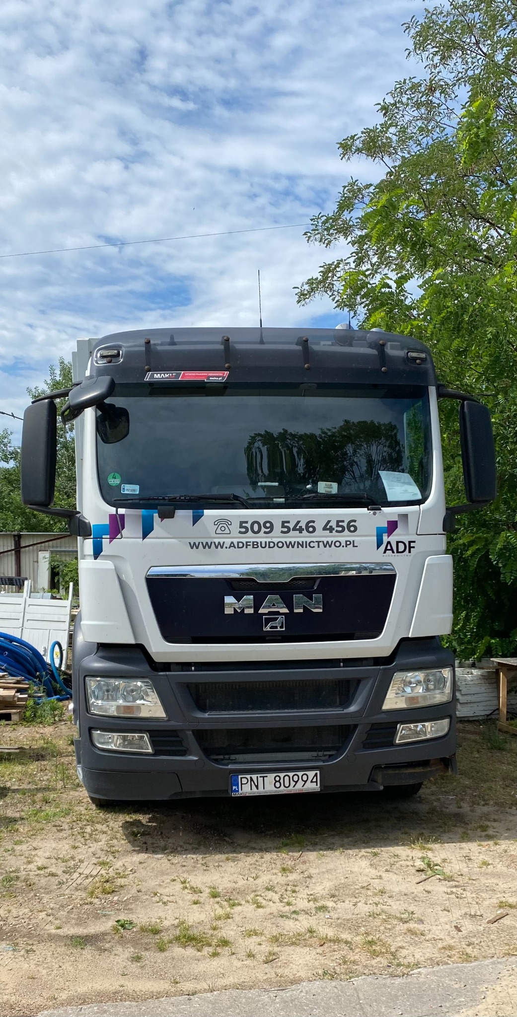 Biała ciężarówka MAN z numerem telefonu i adresem strony internetowej ADF Budownictwo, zaparkowana na piaszczystym terenie.