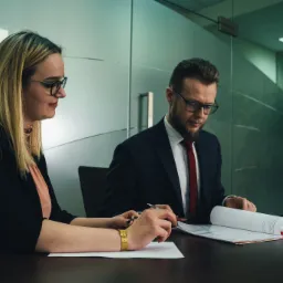 Adwokat i klientka analizują dokumenty przy biurku w nowoczesnym biurze, kobieta w czarnej marynarce i złotej bransoletce, mężczyzna w garniturze z czerwoną kravatą.