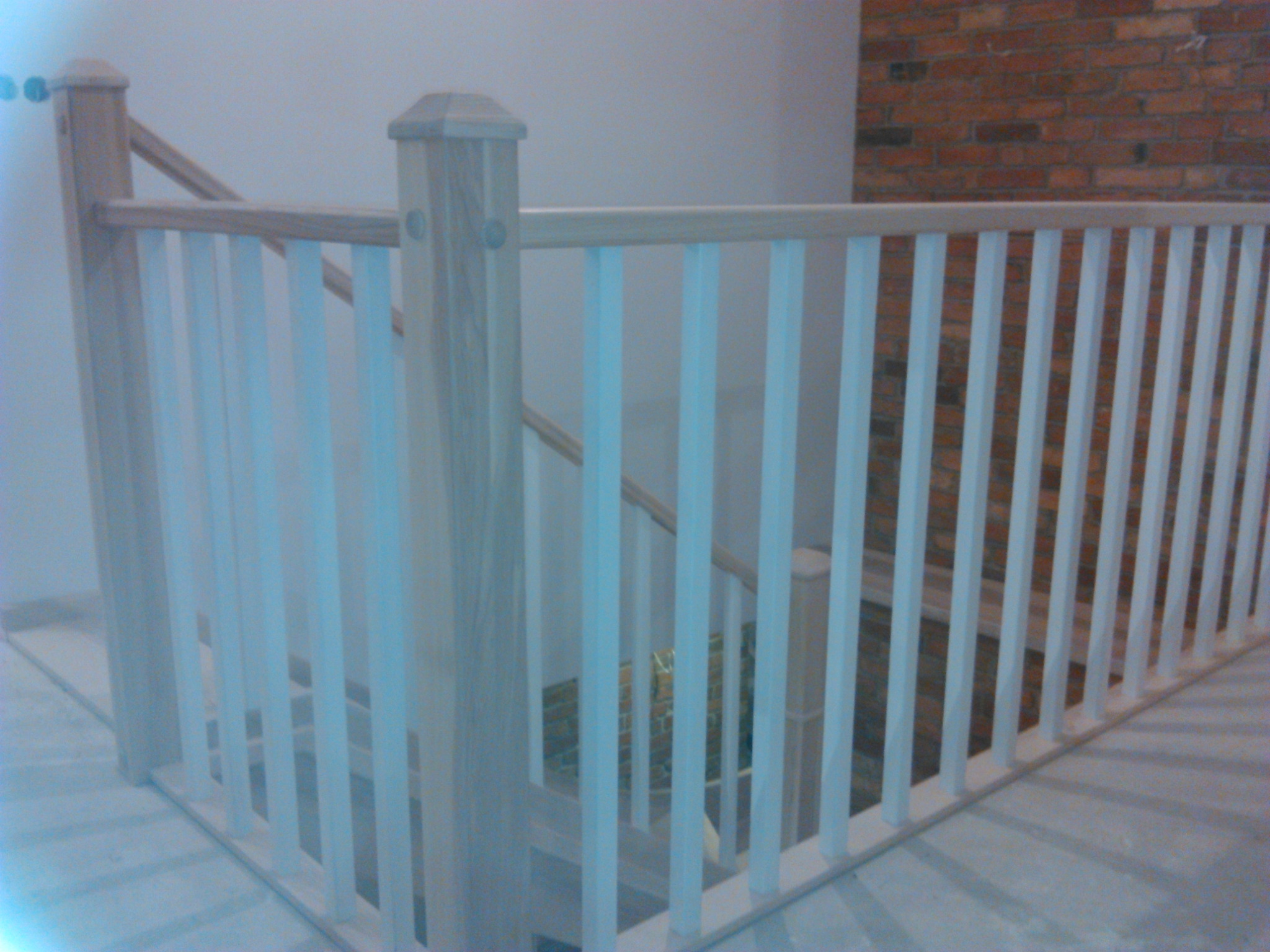 Jasna, drewniana balustrada schodowa z białymi pionowymi szczeblami, widok z góry na schody i ceglaną ścianę w tle.