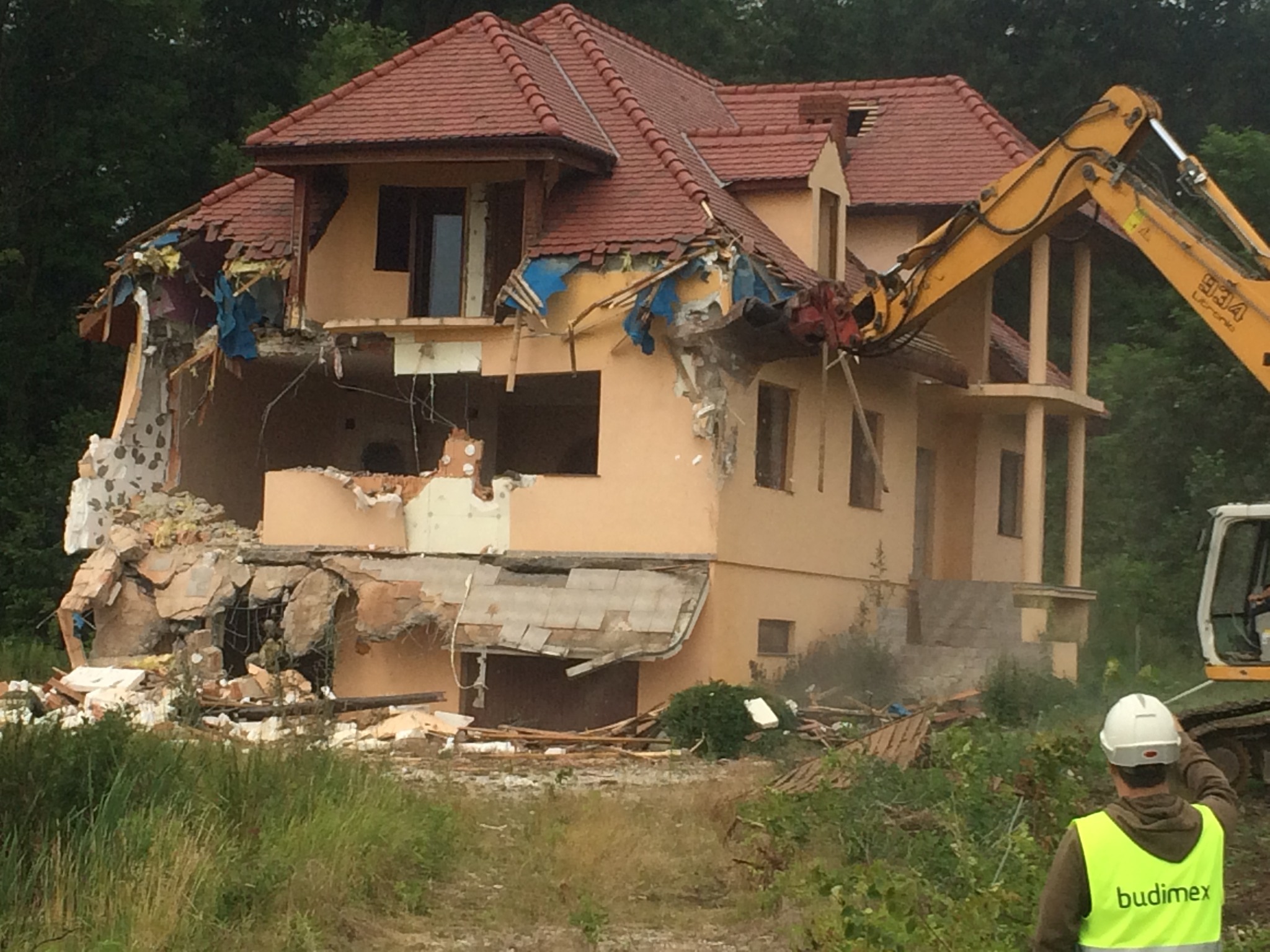 Żółta koparka z hydraulicznym młotem rozbiera dwupiętrowy dom z czerwonym dachem, widoczny pracownik w kasku i kamizelce odblaskowej z logo firmy.