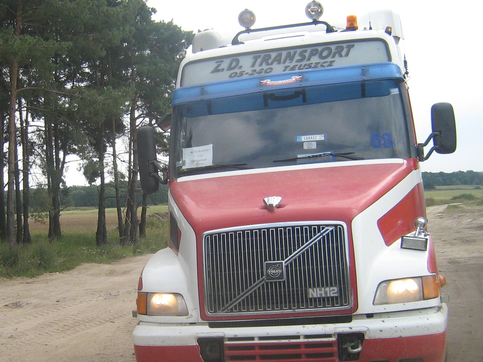 Czerwono-biała ciężarówka Volvo NH12 Z.D. Transport na polnej drodze, widok z przodu, z lasem w tle.