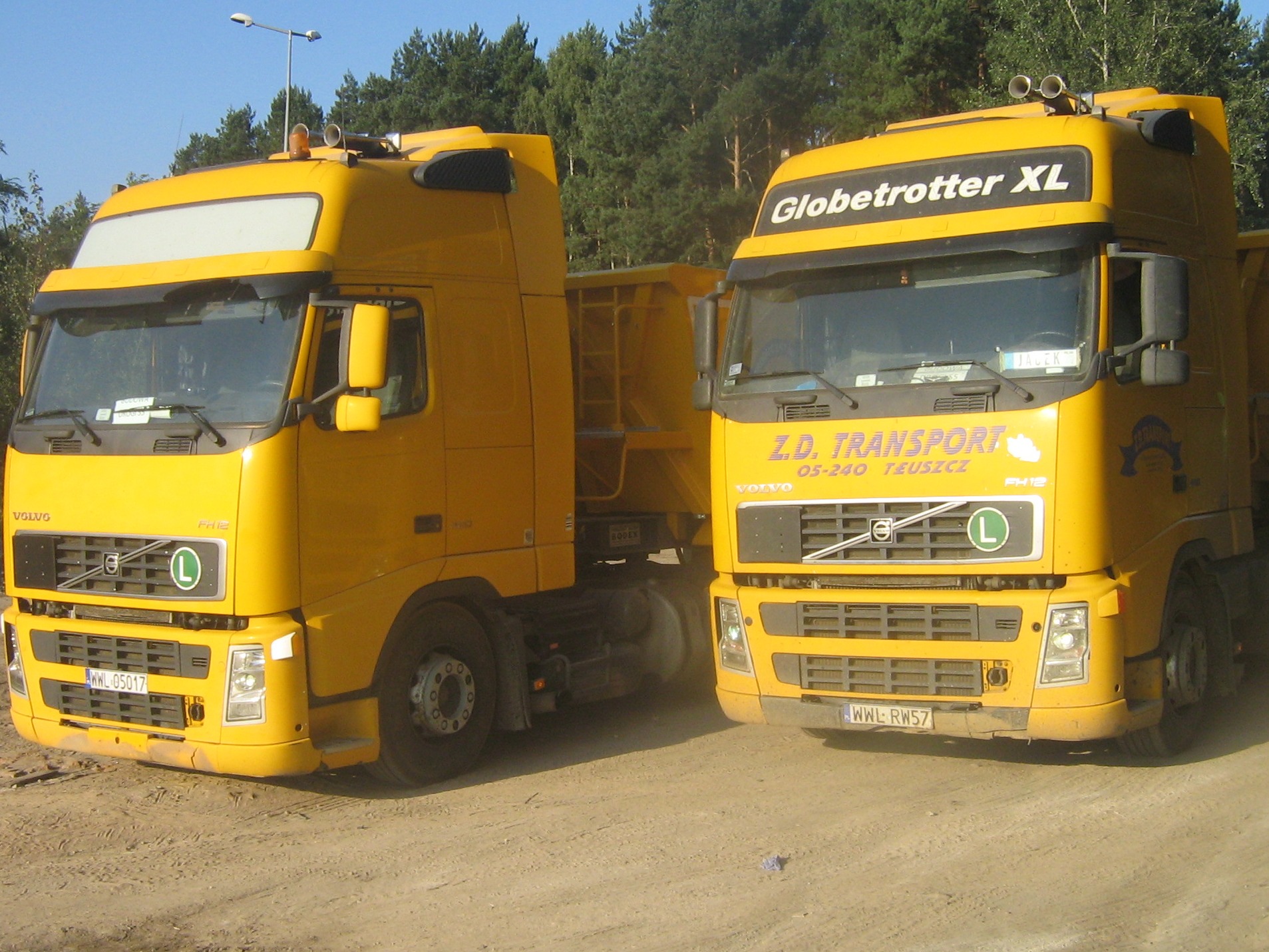 Dwa żółte samochody ciężarowe Volvo Globetrotter XL z naczepami na piaszczystym terenie, prawdopodobnie wykorzystywane do transportu materiałów budowlanych.