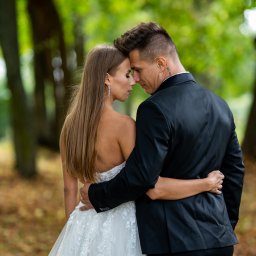 StudioFOTOFILMY - Para młoda, panna młoda w sukni z odkrytymi ramionami i pan młody w czarnym garniturze, obejmują się na tle zielonego parku jesienią, widoczny tatuaż na szyi mężczyzny.