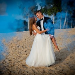StudioFOTOFILMY - Para młoda na plaży w obłokach niebieskiego dymu. Pan młody trzyma pannę młodą na rękach, patrzą sobie w oczy.