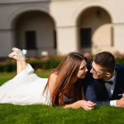 Para młoda leżąca na trawie, panna młoda w białej sukni i srebrnych butach, pan młody w granatowym garniturze z muchą, w tle budynek z arkadami.