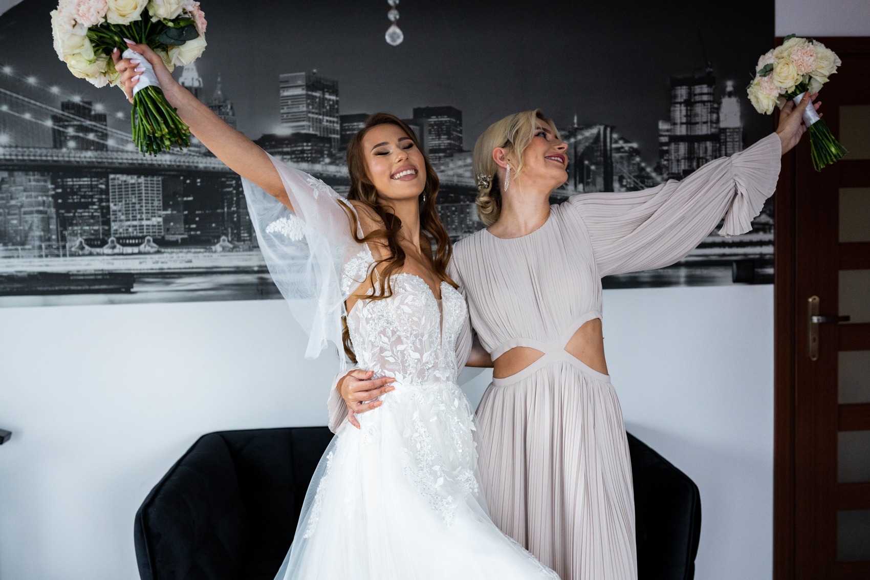 Panna młoda w koronkowej sukni ślubnej i druhna w plisowanej sukience pozują z bukietami na tle czarno-białej fototapety z widokiem na miasto.