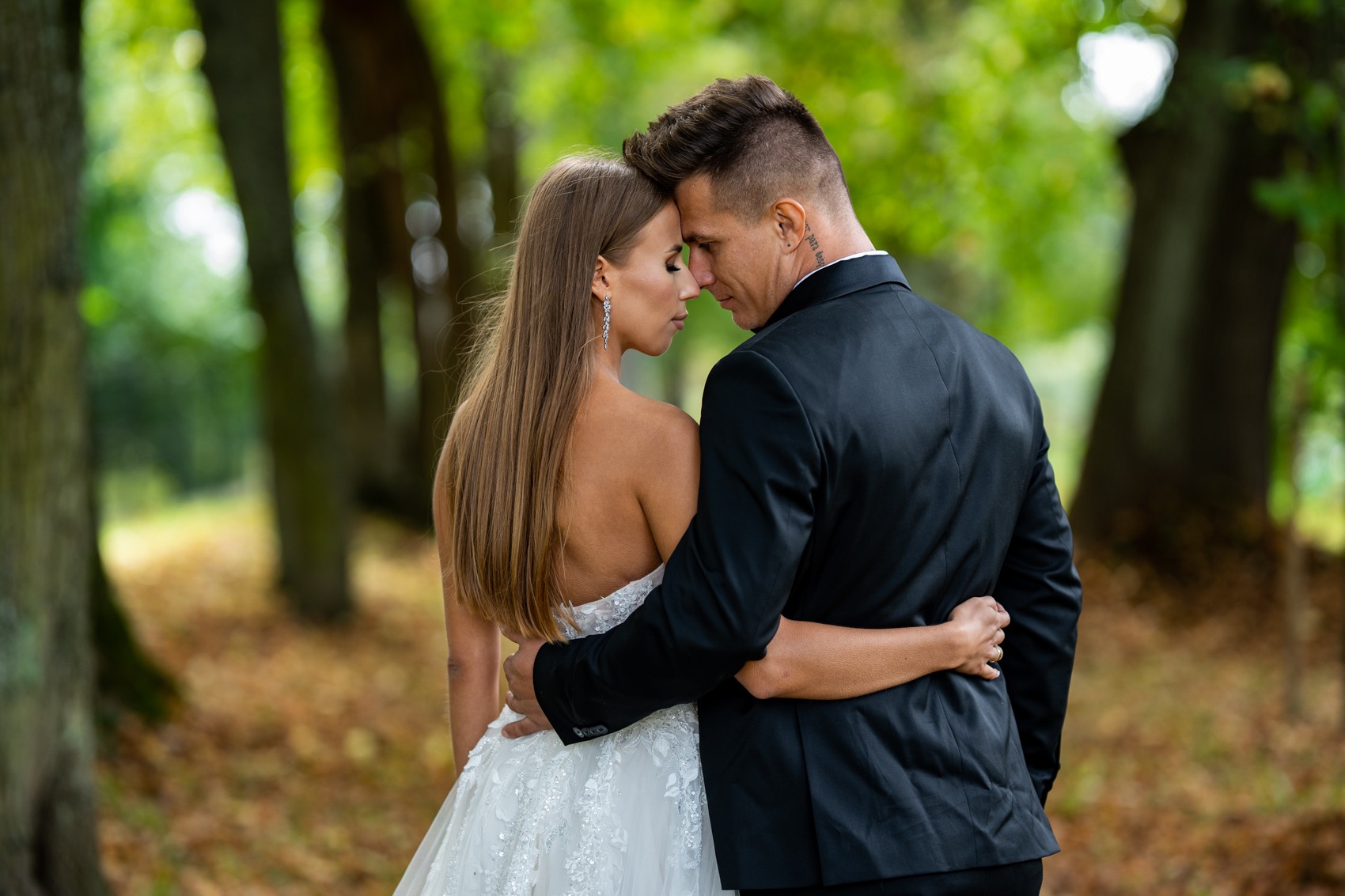 Para młoda, panna młoda w sukni z odkrytymi ramionami i pan młody w czarnym garniturze, obejmują się na tle zielonego parku jesienią, widoczny tatuaż na szyi mężczyzny.