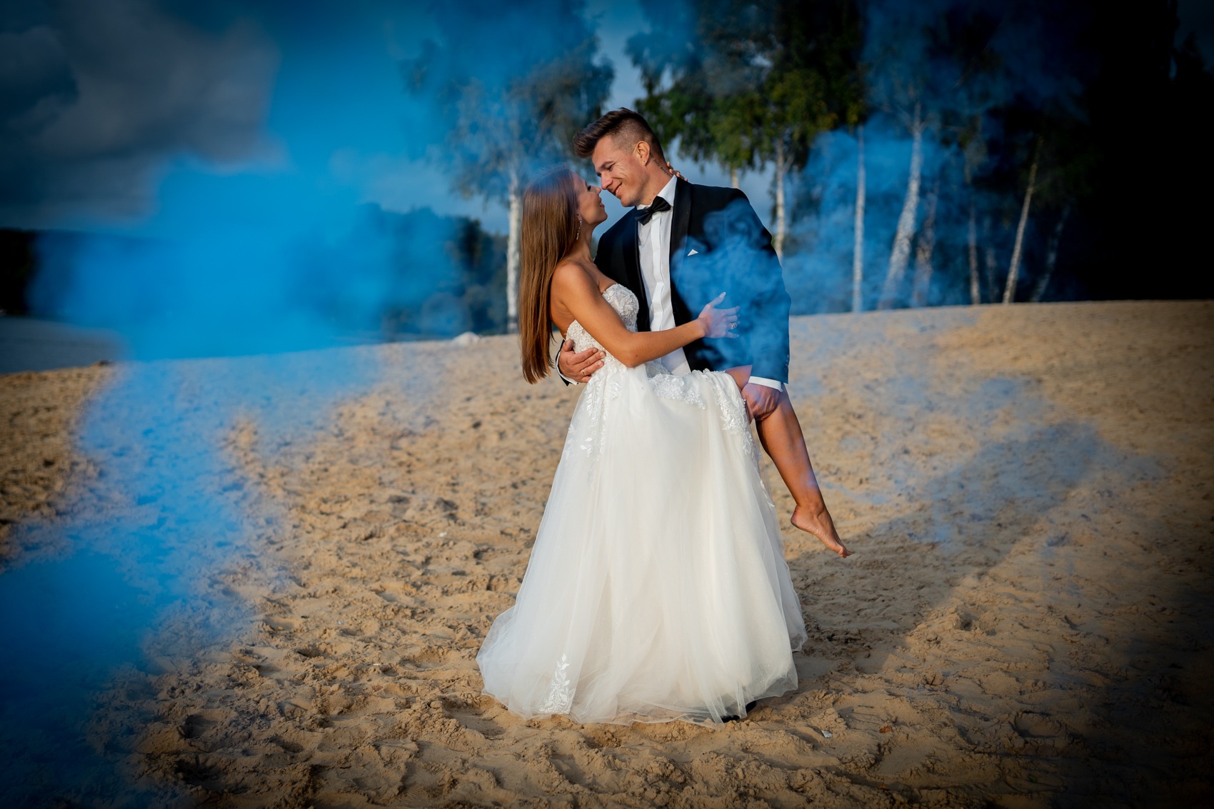 Para młoda na plaży w obłokach niebieskiego dymu. Pan młody trzyma pannę młodą na rękach, patrzą sobie w oczy.