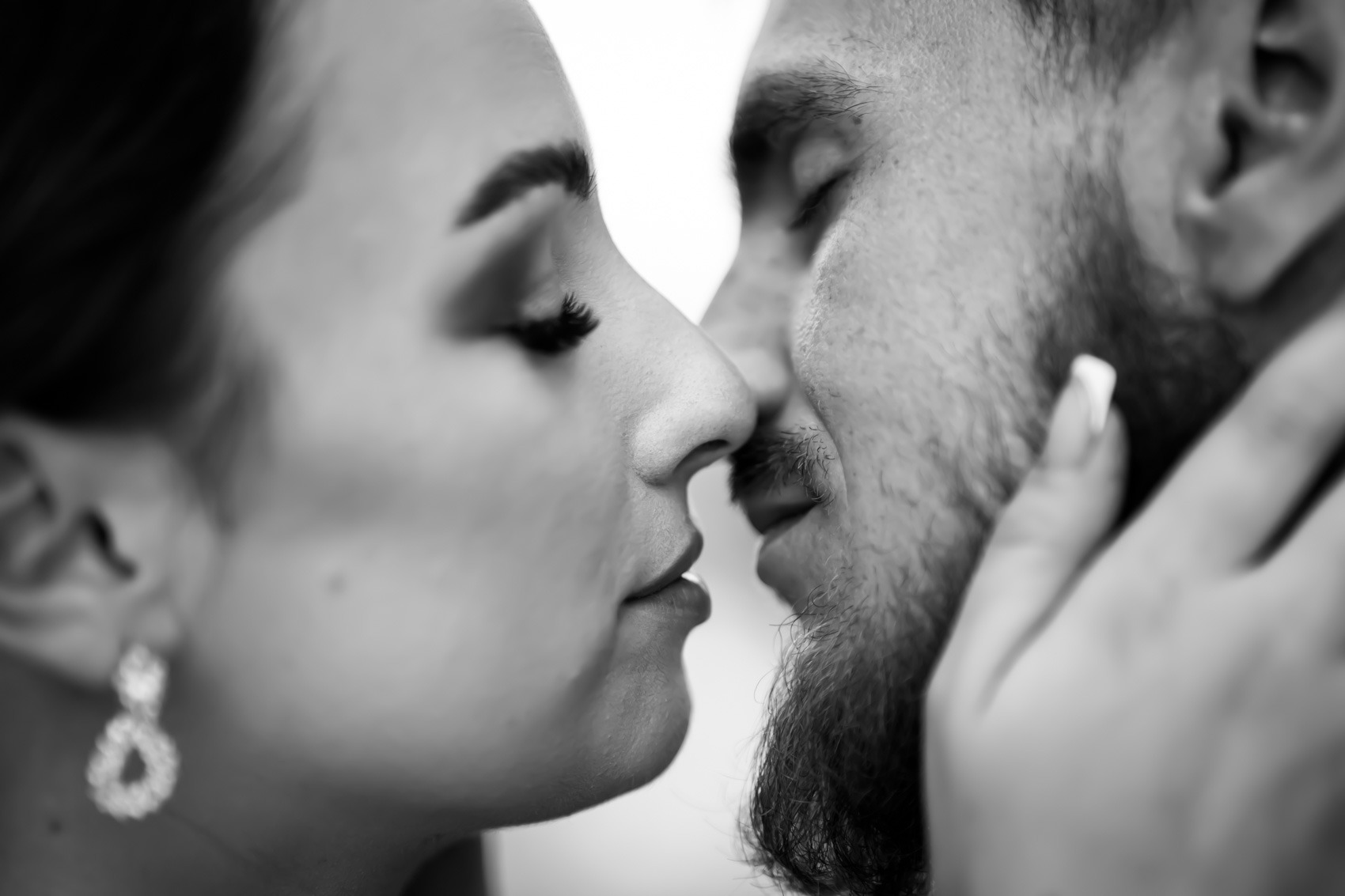 Czarno-białe zbliżenie twarzy pary, z zamkniętymi oczami, delikatnie dotykających się nosami, widoczne kolczyki u kobiety i broda u mężczyzny.