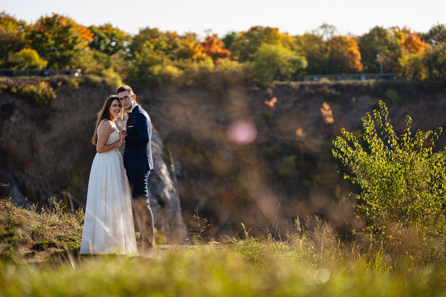 Para młoda w plenerze, ona w białej sukni z brokatem, on w granatowym garniturze, stoją na wzgórzu z widokiem na jesienny las i skalisty klif.