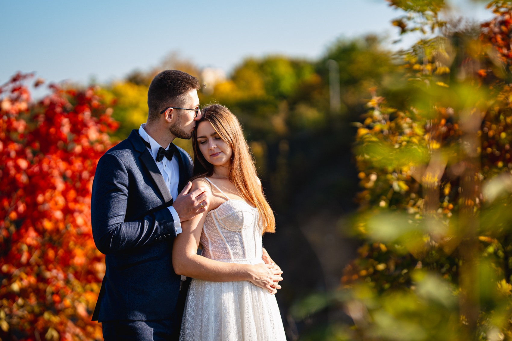 Elegancko ubrana para, mężczyzna w granatowym smokingu z muszką całuje kobietę w brokatowej sukni w czoło, jesienne tło z czerwonymi i żółtymi liśćmi.