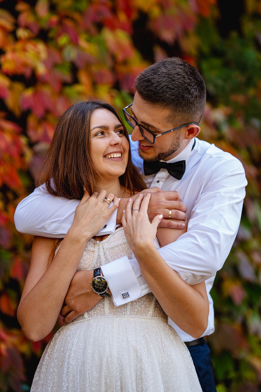 Uśmiechnięta para w objęciach na tle jesiennych liści, ona w jasnej sukience, on w białej koszuli z muchą i mankietami, widoczne obrączki i zegarek.