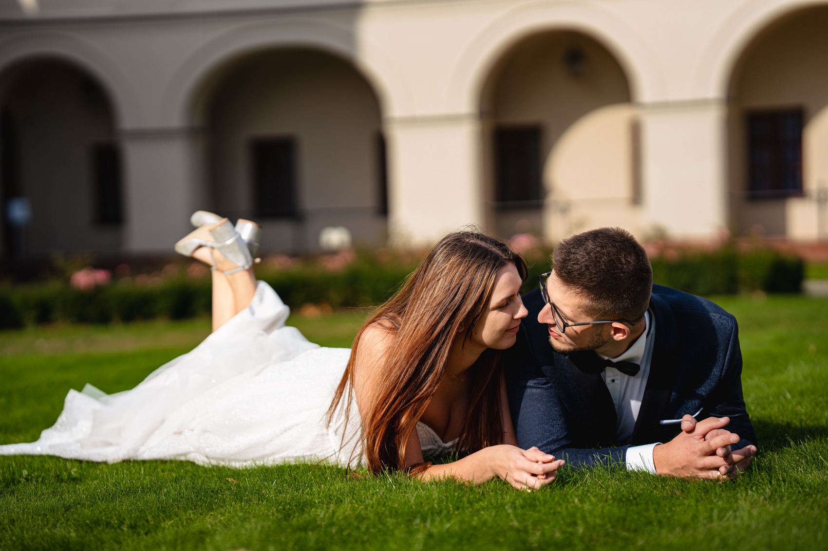 Para młoda leżąca na trawie, panna młoda w białej sukni i srebrnych butach, pan młody w granatowym garniturze z muchą, w tle budynek z arkadami.