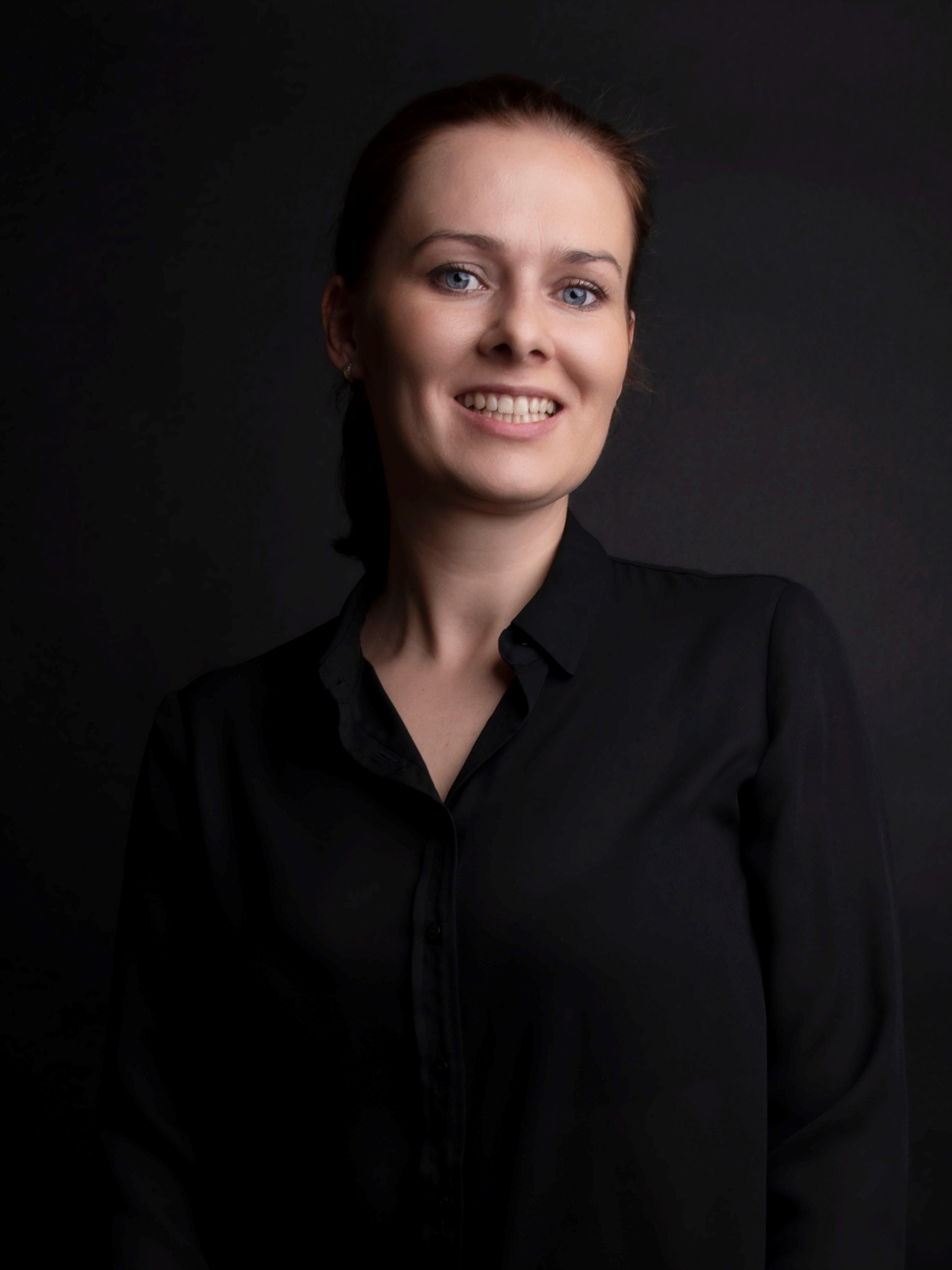 Portret uśmiechniętej kobiety w czarnej koszuli na ciemnym tle, z subtelnymi kolczykami w uszach.
