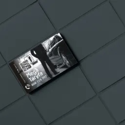 Czarno-biała wizytówka taksówkarska z QR kodem, zdjęciem taksówki i kierowcy, ułożona na ciemnoszarym tle z fakturą.