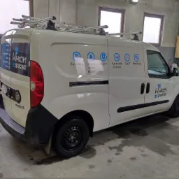 Biały samochód dostawczy z logo firmy Wi-Kam, oferującej usługi instalacji domofonów, anten TV SAT, alarmów, automatyki do bram, sieci IT i monitoringu, zaparkowany w garażu.