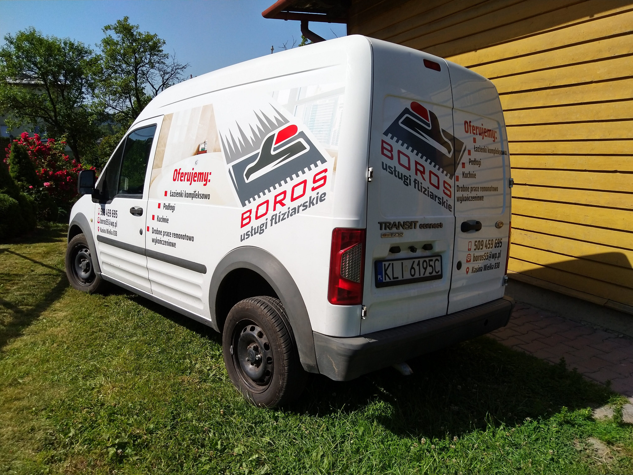 Biały samochód dostawczy z logo firmy świadczącej usługi fliziarskie, zaparkowany na trawie przed drewnianym budynkiem.