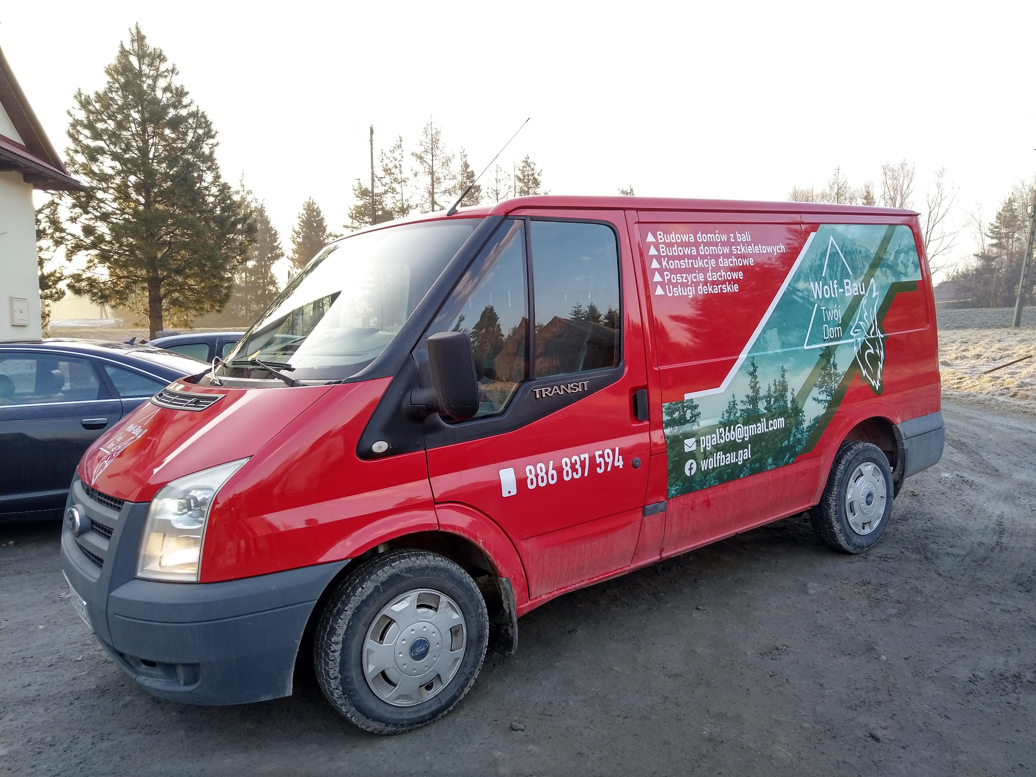 Czerwony samochód dostawczy Ford Transit z oklejeniem reklamowym firmy Wolf-Bau oferującej budowę domów z bali i szkieletowych, konstrukcje dachowe, poszycia dachowe i usługi dekarskie, zaparkowany...