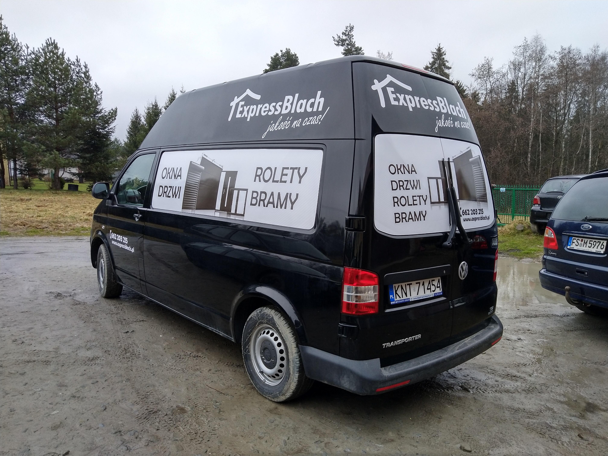Czarny bus firmy ExpressBlach z reklamą okien, drzwi, rolet i bram na karoserii, zaparkowany na błotnistym placu w pochmurny dzień.