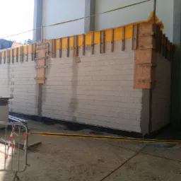 Wznoszenie ściany z bloczków silikatowych z zastosowaniem szalunku i żółtej folii ochronnej na budowie, widoczne materiały budowlane i taśma ostrzegawcza.