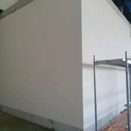 Świeżo pomalowana, jasna ściana w hali przemysłowej z widocznym fragmentem rusztowania i zabezpieczeniem podłoża folią ochronną.