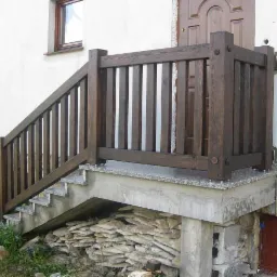 Ciemnobrązowa, drewniana barierka schodowa z pionowymi szczeblami, zamontowana na betonowych schodach z granitowymi płytami, przy białej ścianie budynku z brązowymi drzwiami.