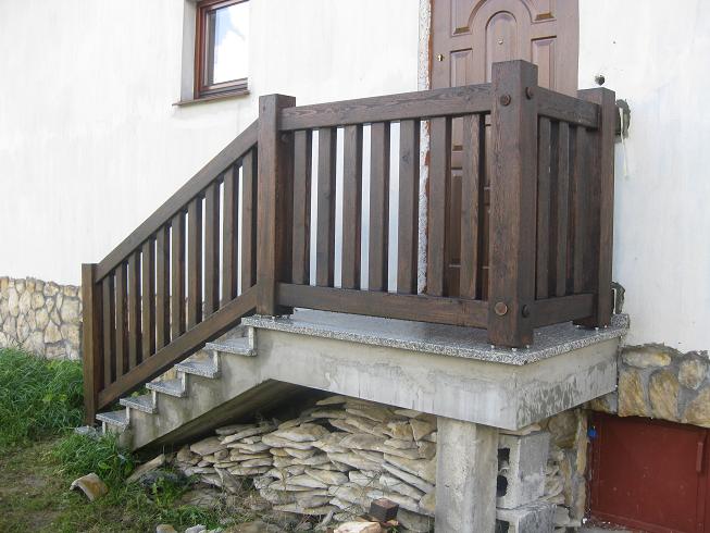 Ciemnobrązowa, drewniana barierka schodowa z pionowymi szczeblami, zamontowana na betonowych schodach z granitowymi płytami, przy białej ścianie budynku z brązowymi drzwiami.