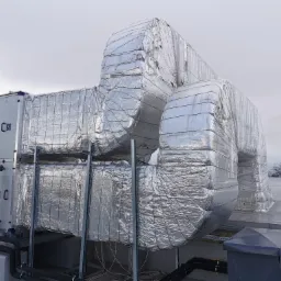 Izolowane kanały wentylacyjne z folii aluminiowej na dachu budynku, widoczny fragment centrali wentylacyjnej i rury w izolacji.