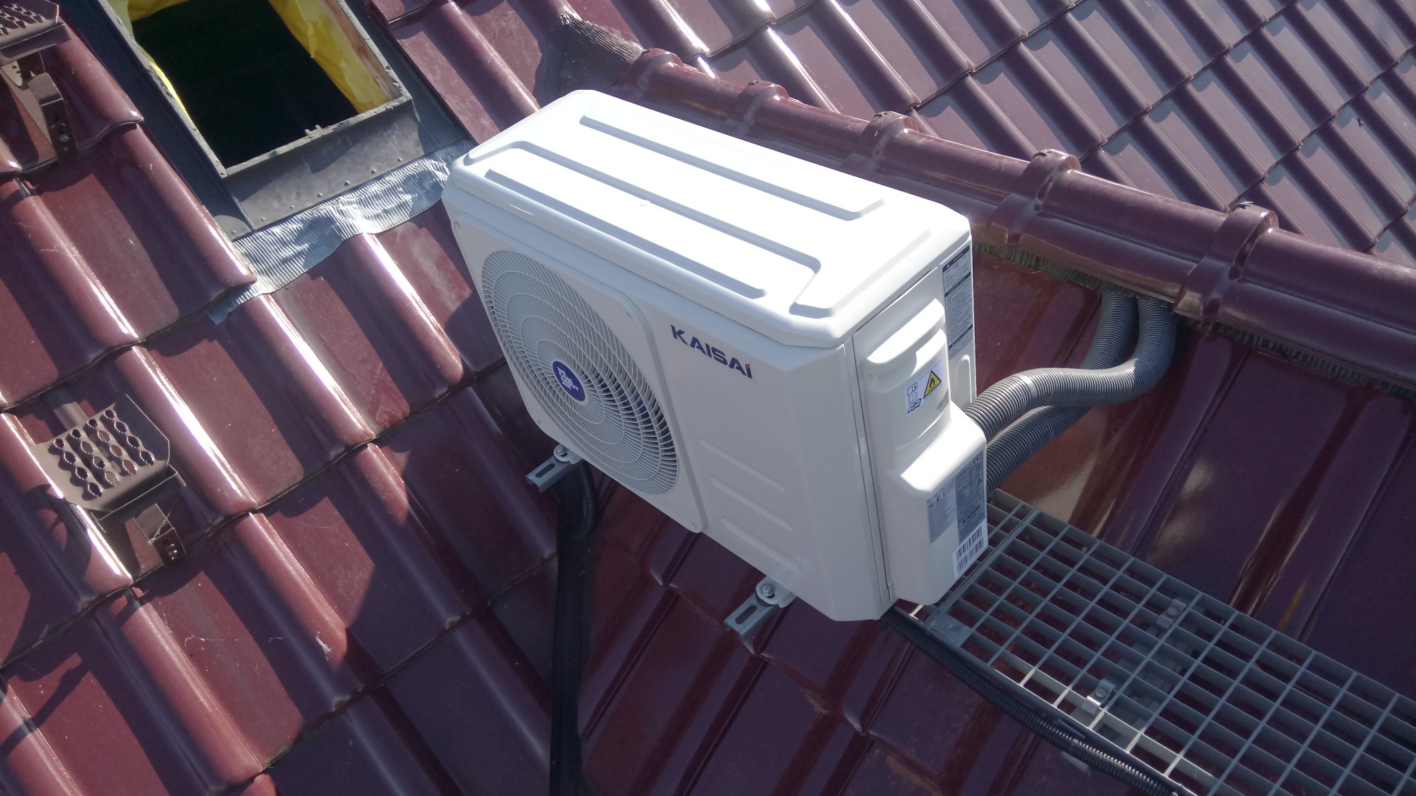 Biała jednostka zewnętrzna klimatyzacji Kaisai zamontowana na brązowym dachu z dachówki ceramicznej, widoczna krata pomostowa i rury instalacyjne.