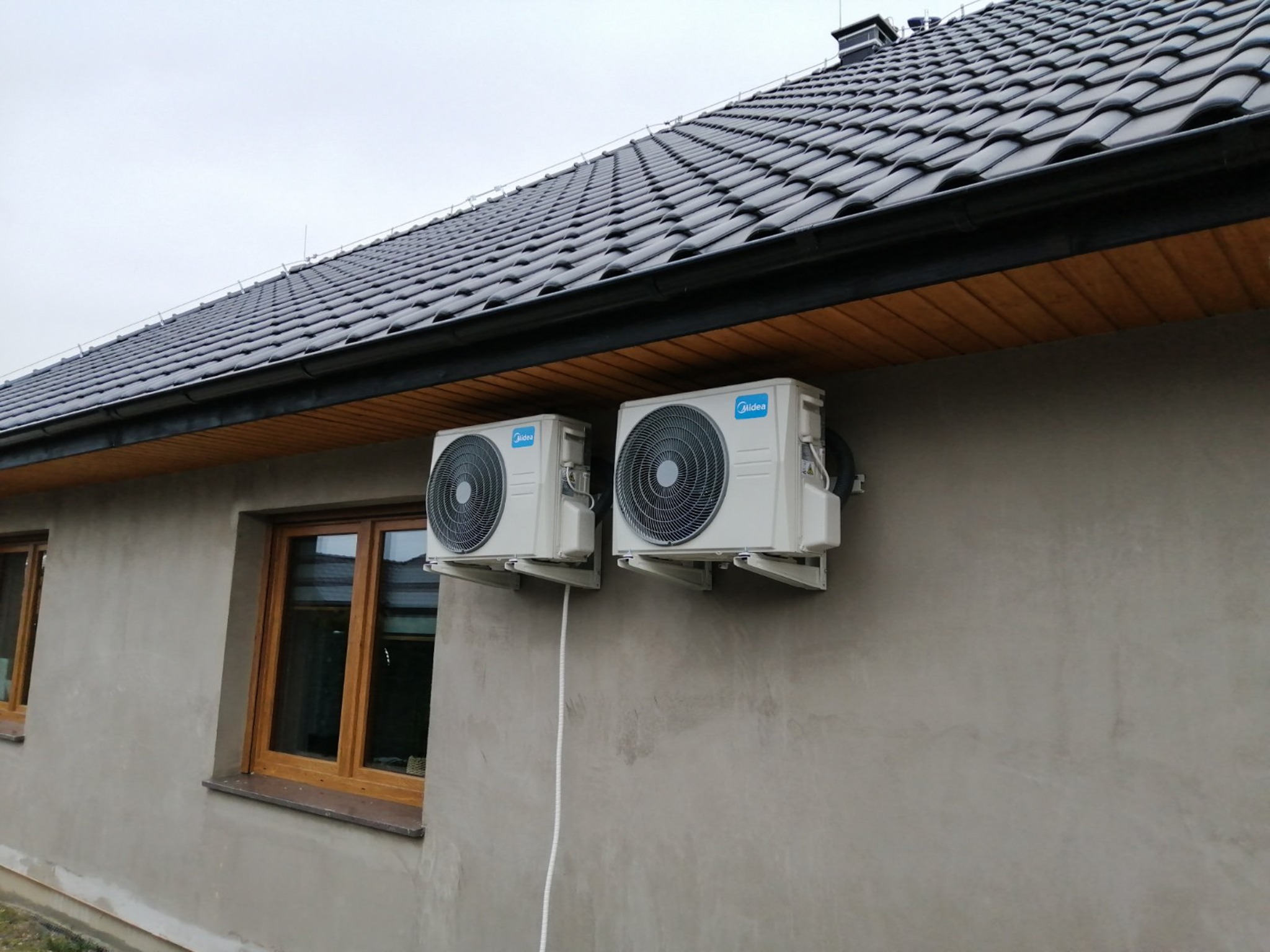 Dwa białe klimatyzatory Midea zamontowane na szarej ścianie domu w Legnicy, widoczne okna z drewnianymi ramami i ciemny dach z rynną.