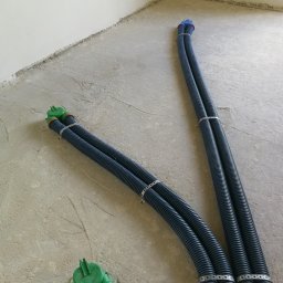 Dom z Energią Sp. z o.o. - Dwie pary karbowanych rur wentylacyjnych, spięte metalowymi opaskami, wychodzące z podłogi w surowym stanie, z zielonymi i niebieskimi zakończeniami.