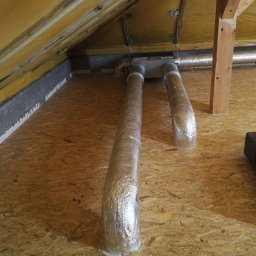 Dom z Energią Sp. z o.o. - Dwie rury wentylacyjne owinięte folią aluminiową, wychodzące z sufitu na poddaszu, zamontowane w drewnianej konstrukcji, z widoczną izolacją i elementami mocującymi.