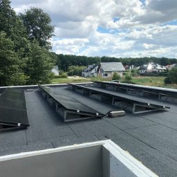 Dom z Energią Sp. z o.o. - Panele słoneczne zamontowane na płaskim dachu pokrytym papą, widok z góry na instalację fotowoltaiczną, w tle domy jednorodzinne i zieleń.
