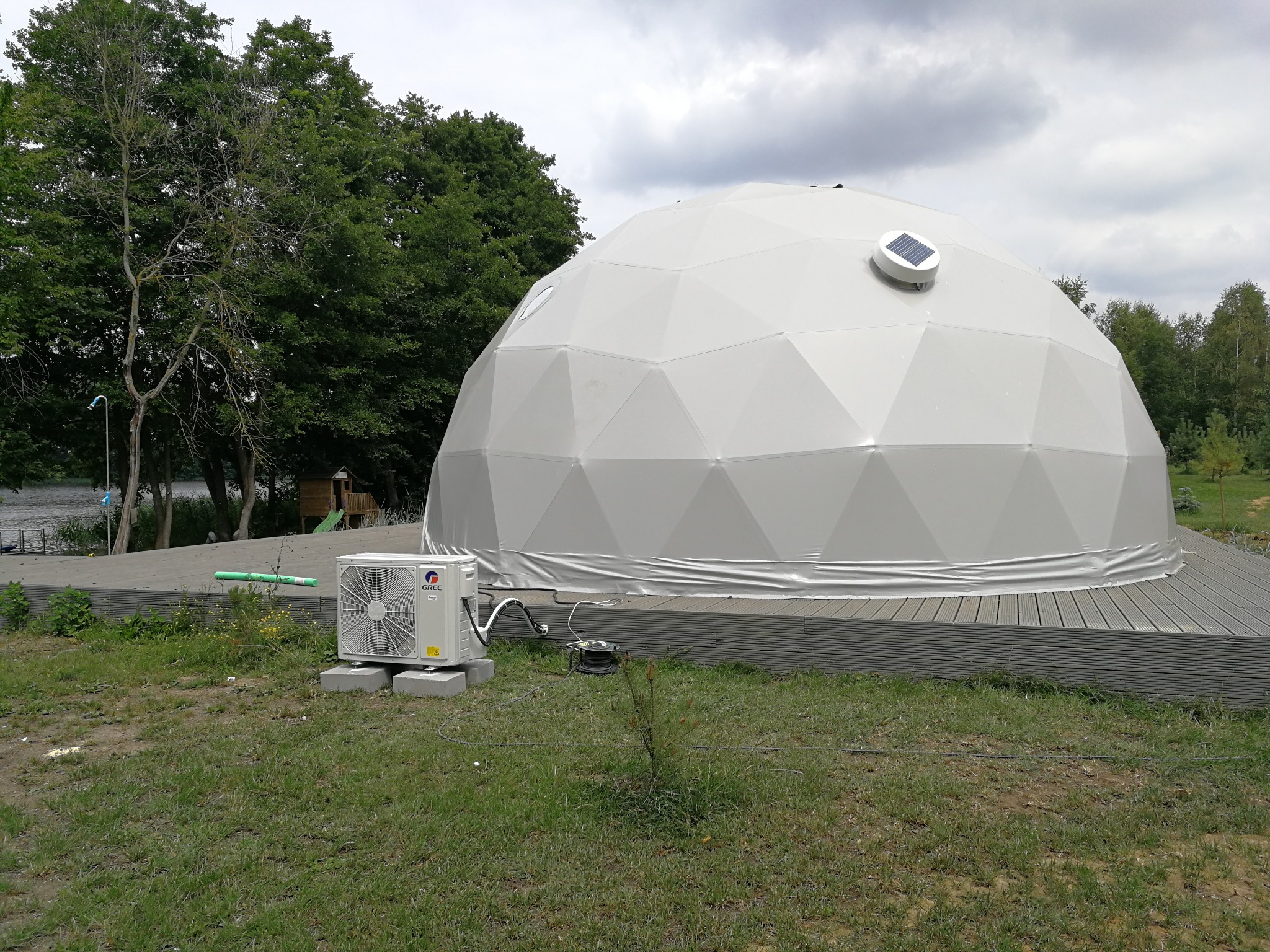 Biała geodezyjna kopuła z panelem solarnym na szczycie, umieszczona na drewnianym tarasie, z jednostką zewnętrzną klimatyzacji marki Gree stojącą na trawie obok.