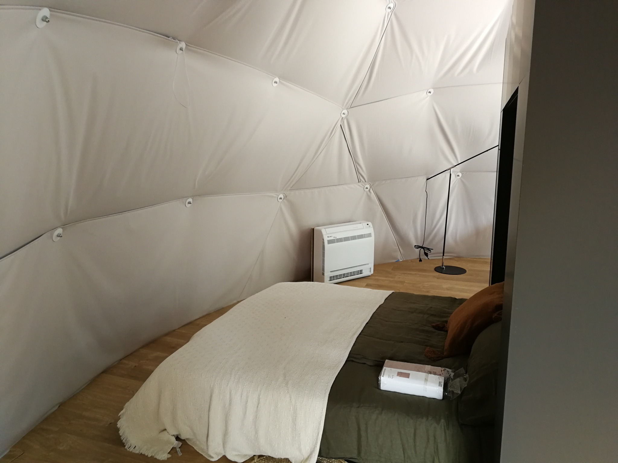 Wnętrze kopuły geodezyjnej z białym, tekstylnym pokryciem, wyposażone w łóżko z zieloną pościelą, białą narzutą, brązową poduszką i białą jednostką ścienną, prawdopodobnie grzewczą, oraz czarną...