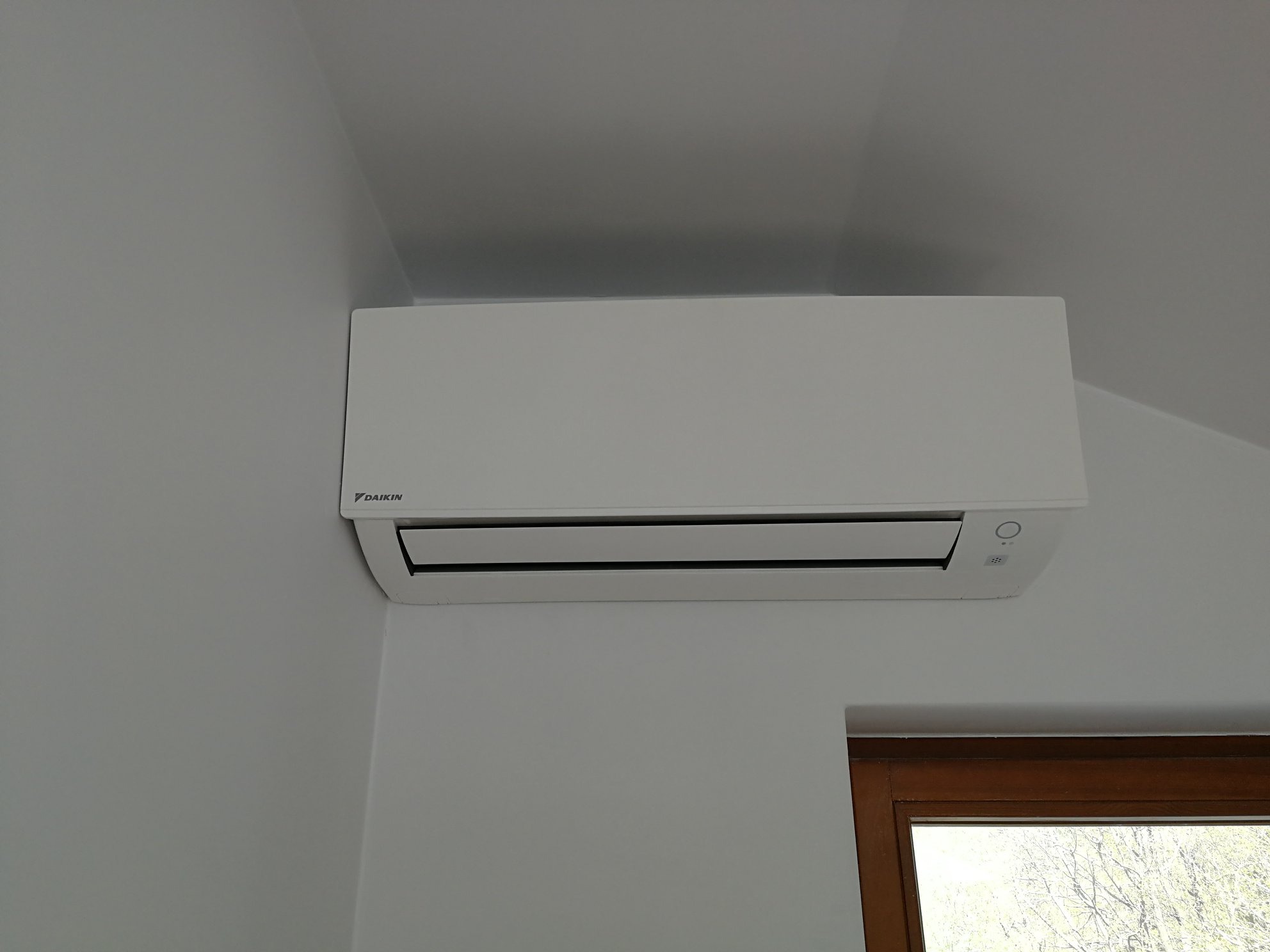 Biała jednostka wewnętrzna klimatyzatora Daikin zamontowana na ścianie, widoczne fragmenty ramy okiennej w prawym dolnym rogu.