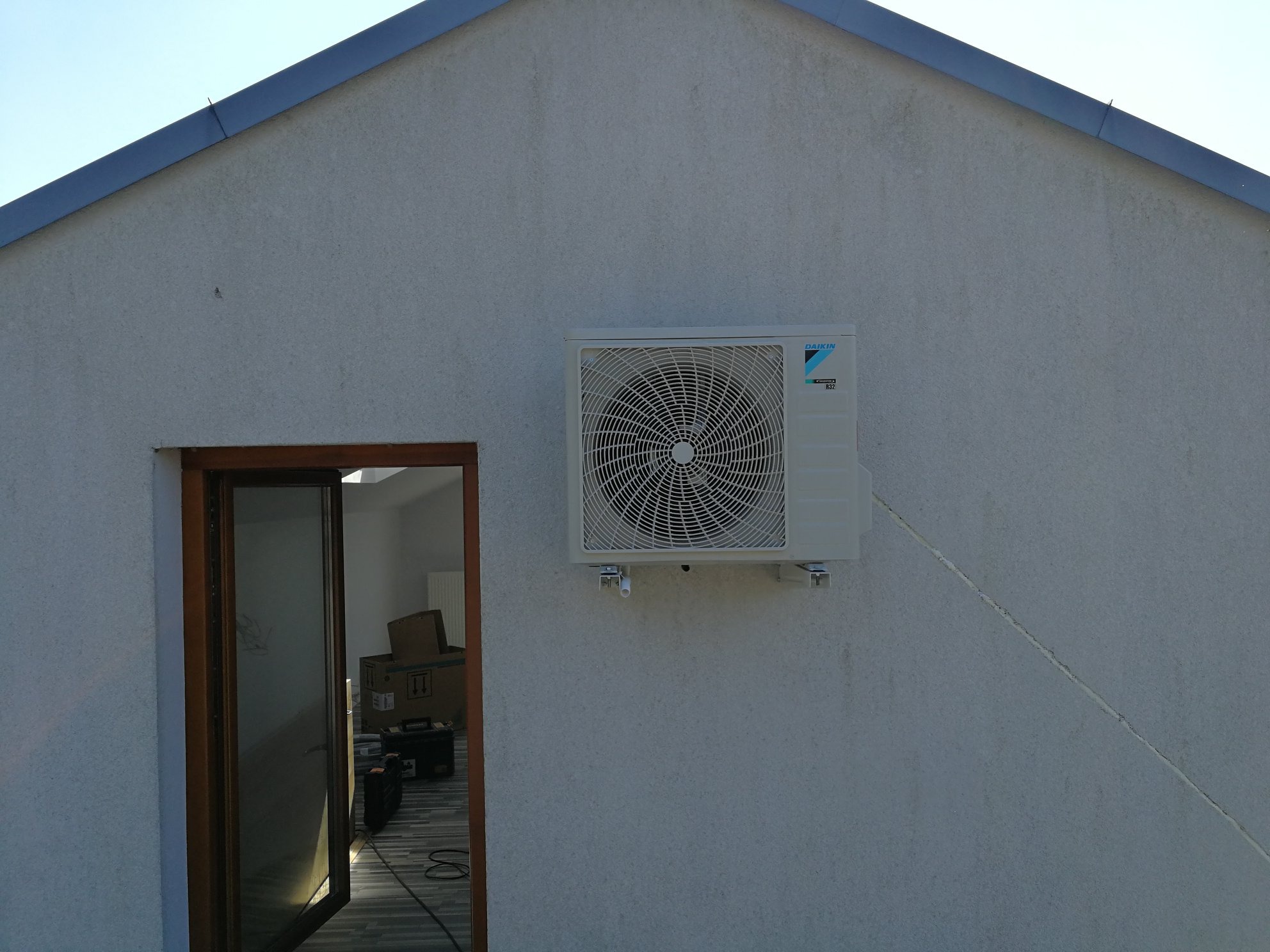 Biała jednostka zewnętrzna klimatyzacji Daikin zamontowana na jasnej, teksturowanej ścianie budynku z widocznym oknem i elementami wyposażenia wnętrza w tle.