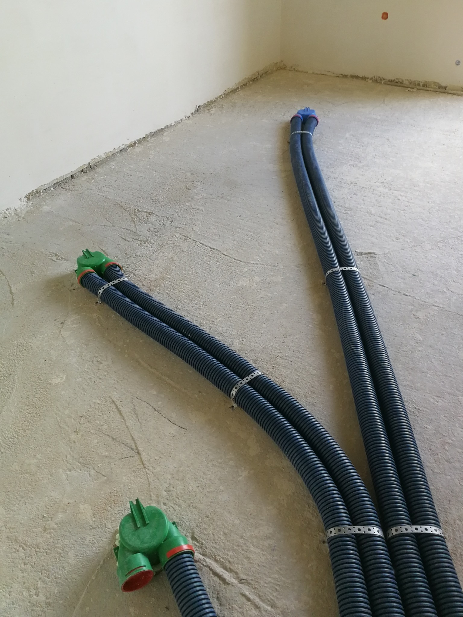 Dwie pary karbowanych rur wentylacyjnych, spięte metalowymi opaskami, wychodzące z podłogi w surowym stanie, z zielonymi i niebieskimi zakończeniami.
