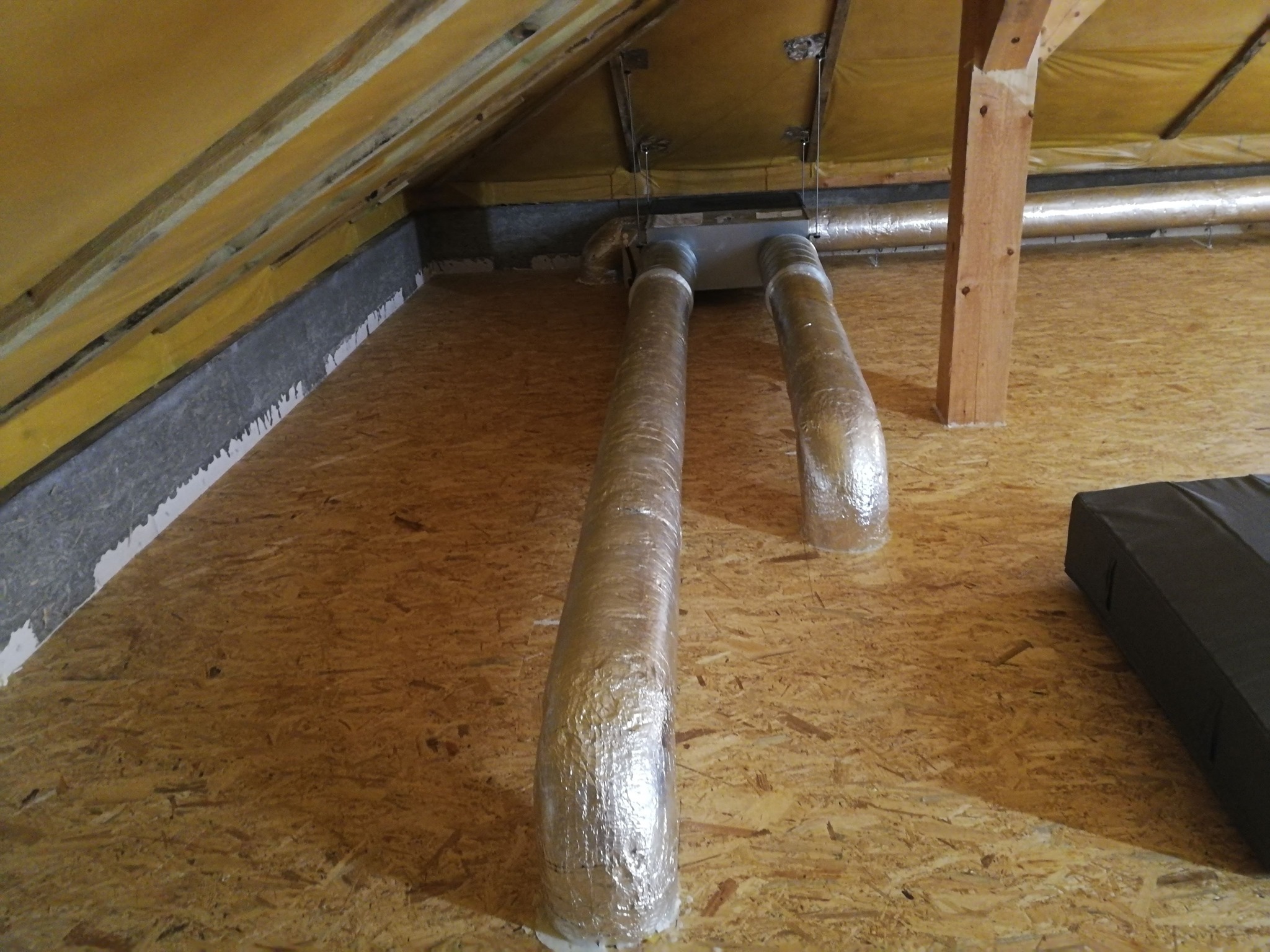 Dwie rury wentylacyjne owinięte folią aluminiową, wychodzące z sufitu na poddaszu, zamontowane w drewnianej konstrukcji, z widoczną izolacją i elementami mocującymi.