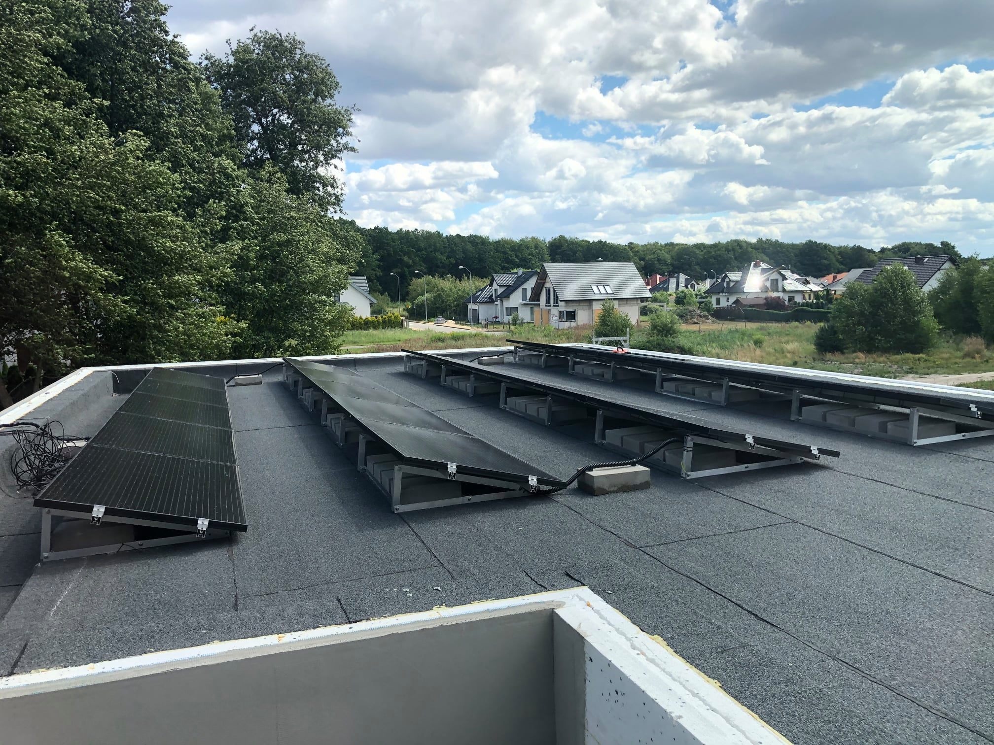 Panele słoneczne zamontowane na płaskim dachu pokrytym papą, widok z góry na instalację fotowoltaiczną, w tle domy jednorodzinne i zieleń.