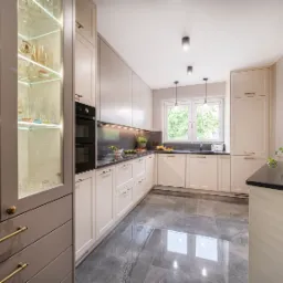 Jasna kuchnia z szarymi marmurowymi płytkami podłogowymi, przeszklona szafka z podświetlonymi szklankami i butelkami, czarny blat, ceglana ściana z dekoracją, okno z widokiem na zieleń.