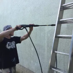 Mężczyzna w czarnej koszulce z tatuażem na ramieniu, stojący na drabinie, czyści elewację domu myjką ciśnieniową.