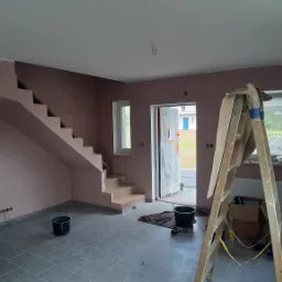 Wnętrze w trakcie prac: świeżo pomalowane na różowo ściany, drewniana drabina oparta o ścianę pod oknem, wiaderka z farbą na podłodze z szarych płytek, widoczne schody.