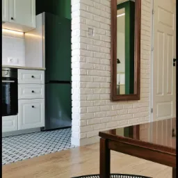 Nowoczesne wnętrze: biała kuchnia z czarnymi akcentami, ceglana ściana z lustrem w drewnianej ramie, podłoga z wzorzystymi płytkami przechodząca w drewniany parkiet z okrągłym dywanem.