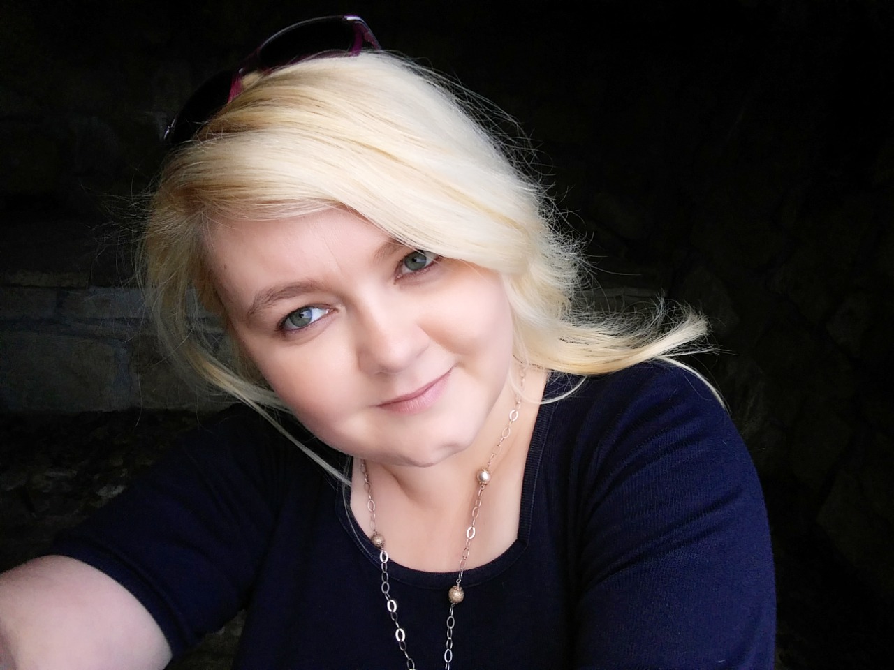 Portret uśmiechniętej blondynki z okularami na głowie, w ciemnej bluzce i z delikatnym naszyjnikiem, na tle ciemnej ściany z kamienia.