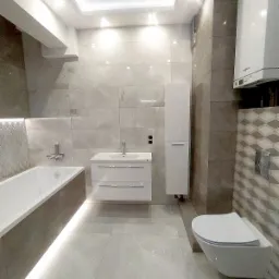 Nowoczesna łazienka z podświetlaną wanną, wiszącą szafką umywalkową, toaletą i grzejnikiem, wykończona szarymi płytkami w różnych odcieniach.