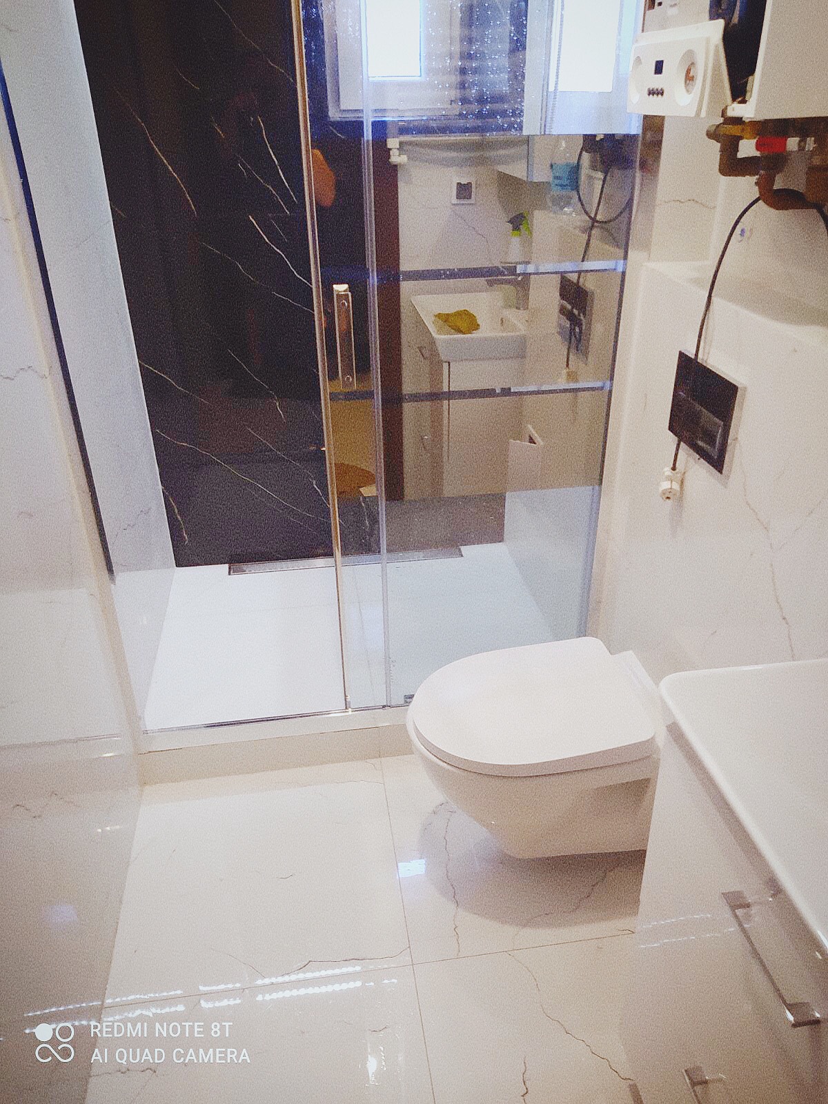 Nowoczesna łazienka z kabiną prysznicową typu walk-in z czarnym panelem, podwieszaną toaletą i umywalką z szafką, wykończona białymi płytkami z szarymi żyłami imitującymi marmur.